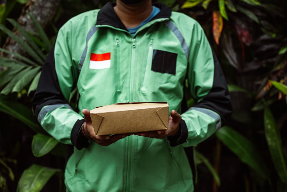 A delivery person wearing a bright green jacket with black sleeves, a black zip-up front, and a red-and-white flag patch on the chest is holding a plain, rectangular cardboard box with both hands. The
