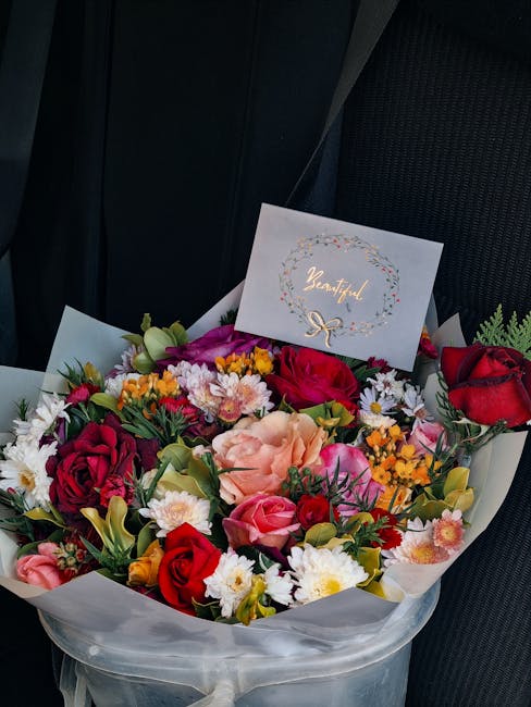 A vibrant floral arrangement featuring a variety of fresh flowers including red roses, pink roses, peach carnations, white daisies, purple and yellow blooms, and green foliage. The bouquet is wrapped