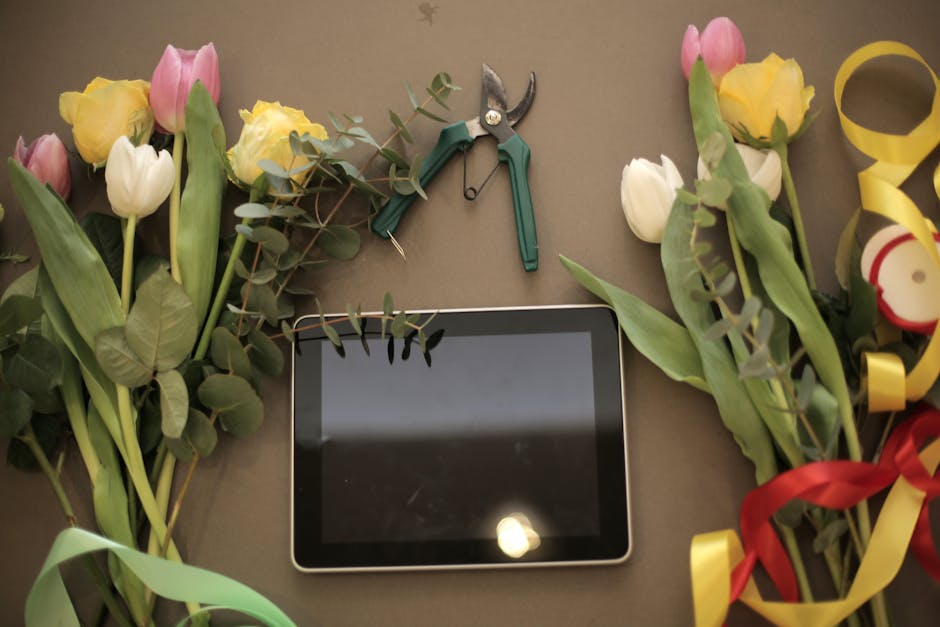 A flat lay image featuring fresh tulips in shades of pink, white, and yellow, arranged with eucalyptus leaves and greenery. The flowers are partially wrapped with decorative ribbons in yellow and red,