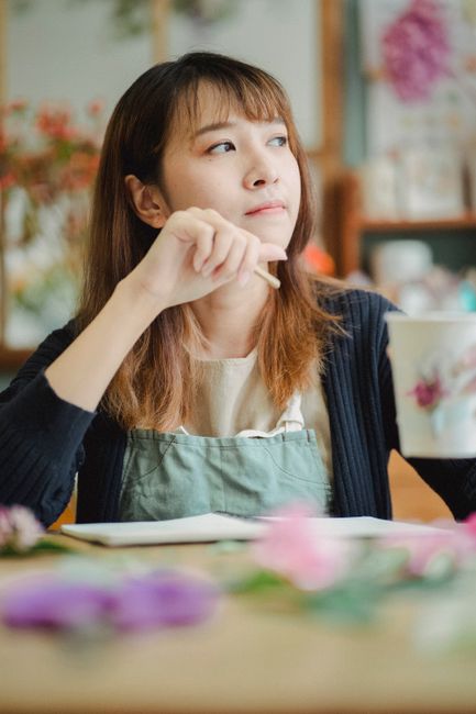 A close-up of a woman holding a pair of scissors, arranging a bouquet of fresh flowers. The arrangement features a vibrant mix of pink, purple, and white blooms with green foliage, wrapped in pastel-c