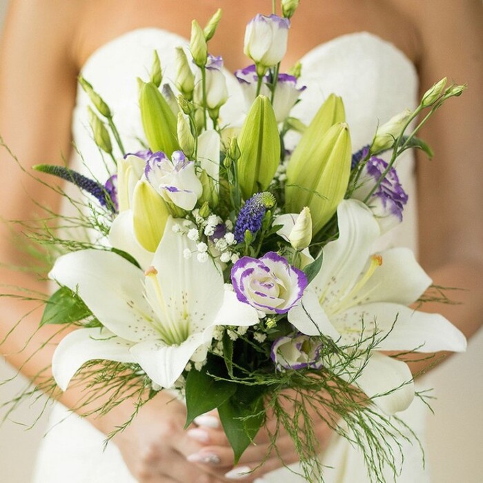 A captivating close-up reveals a South Norwood bride holding an exquisite, locally inspired wedding bouquet. This elegant hand-tied design is a harmonious mix of large, open white Oriental lilies whose scented, speckled petals and vivid yellow-orange stamens evoke a serene sophistication perfect for nearby venues or garden ceremonies. Between the lilies cluster white lisianthus blooms, their ruffled petals accentuated by graceful lavender-purple edging-a nod to the gentle colours of early summer mornings in South Norwood's green spaces. Deep indigo-purple Veronica springs add depth, rising with a quiet drama, while delicate clouds of baby's breath offer a soft, ethereal frame to the arrangement. Unopened lily buds and broad greens infuse a sense of promise and freshness, while airy asparagus fern dances along the bouquet's periphery. The bride's hands, meticulously groomed, support the stems with quiet pride, her classic white strapless dress a blurred yet inviting background. This floral composition, expertly crafted by South Norwood's florists and reminiscent of nearby South Norwood Country Park, captures the essence of romance, purity, and heartfelt celebration-an ideal centrepiece for any South London wedding, blending local heritage and contemporary elegance in every petal.