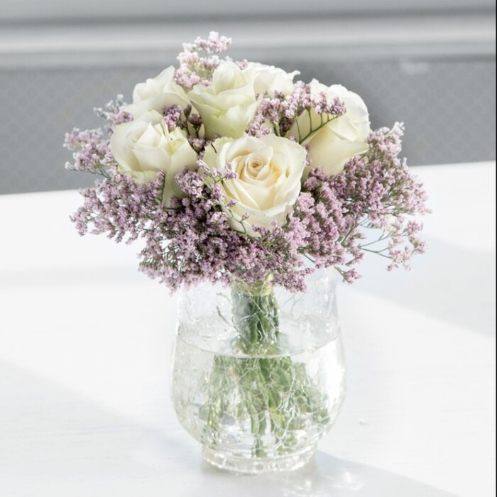At the centre of the composition sits a carefully crafted, domed bouquet of creamy white roses interlaced with clouds of soft lavender statice, the sort of piece a devoted florist in South Norwood would prepare for a heartfelt occasion. The roses are plentiful, around a dozen blooms, each one full and gently opening, with outer petals curving gracefully away from the heart of the flower. Their colour leans toward warm ivory, not a cold white, giving them a luminous quality that suggests quiet affection and timeless elegance. Nestled among these larger blooms, the lavender statice forms a fine, lace-like network of tiny blossoms. Its light purple hue is muted and soothing rather than bright, offering a whisper of colour that softens the structured form of the roses. Together, they create a romantic, tranquil atmosphere, perfect for a sympathy gesture, an anniversary in a stylish South Norwood townhouse, or simply to bring calm to a home office overlooking High Street. The stems are gathered with invisible care and placed in a tall, clear cylindrical glass vase. Through the glass, you can see the straight, green stems overlapping and disappearing into clear water, a reassurance of freshness and recent preparation. The vase stands on a pristine white surface that mirrors a hint of the bouquet's shape, subtly amplifying its presence without distracting from it. Behind, the background is out of focus, washed in soft daylight that appears to stream through a large window, similar to those in modern flats near Norwood Junction. This diffused light touches the rose petals and statice tips, revealing delicate textures and faint shadows. The overall feeling is one of composed serenity and thoughtful design, as though the bouquet has been placed in a quiet room to honour memories, celebrate enduring love, or simply lend a note of gentle, living beauty to an otherwise still space.