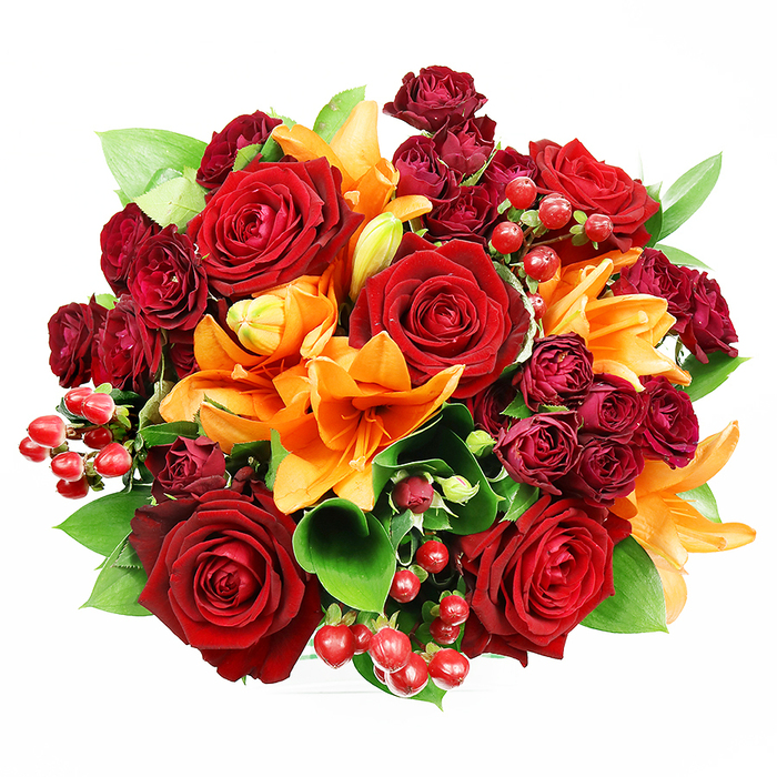 Viewed from above on a spotless white field, this opulent, circular bouquet presents an eloquent mix of texture, colour, and florist skill. Large, fully expanded deep-red roses form the visual heart, their velveteen petals opening in layered spirals that read as both romantic and sumptuous. Around and between them, bright orange Asiatic lilies introduce strong architectural shape - trumpet-like petals, occasional dark freckles, and exposed stamens on the most open blooms - while other lily buds hold the suggestion of further unfolding. Interwoven are tight clusters of dark red or burgundy spray roses whose miniature faces add concentrated, velvety depth and a luxurious contrast to the fuller roses. Small clusters of glossy red hypericum berries are placed like tiny gems, their round, reflective surfaces offering lively highlights and tactile interest among the petals. Cool green foliage - broad, glossy leaves consistent with ruscus or salal - peeks through the composition, providing structural support and a fresh, verdant frame that makes the warm reds and oranges sing. The soft, even lighting reveals the delicate sheen of petal surfaces, the berries' glossy reflections, and subtle shadowing that gives the bouquet dimensionality without drama. Imagining the bouquet in person suggests a layered scent profile of deep rose warmth balanced by the lily's brighter, spicy notes. This arrangement, with its autumnal red-orange-green harmony, feels right for anniversaries, romantic gestures, or celebratory displays and would sit beautifully on a dining table or mantel in a home near South Norwood Lake or along Whitehorse Lane.