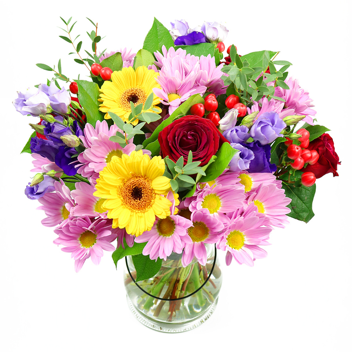 A tall, rounded bouquet rests in a clear glass vase, set against a pure white background that allows every colour to glow with clarity, much like flowers catching soft daylight through a South Norwood bay window. Deep red roses form the emotional core of the design, their layered, velvety petals opening in perfect spirals that evoke enduring affection. Nearby, yellow gerbera daisies with dark brown-orange centres act like bright beacons, their flat, daisy-like faces bringing a joyful, almost playful tone, as if echoing the friendly bustle around Norwood Junction. Encircling these focal blooms, clusters of pink daisies or chrysanthemums create a delicate pastel halo. Each blossom offers a multitude of fine petals radiating from a tiny golden centre, adding both volume and a soft, comforting texture, reminiscent of cottage gardens hidden along side streets off High Street. Interlaced through this gentle pink cloud, purple and lavender lisianthus blooms introduce cooler tones. Their bell-shaped flowers, with slightly crimped and curled edges, lend a sense of movement and refinement, as though the bouquet has been styled for a special occasion dinner near South Norwood Country Park. Scattered pockets of bright red Hypericum berries appear like tiny, polished beads, giving the arrangement a distinctive modern twist and adding interest from every viewing angle. The design is completed with layers of greenery in varied shapes and hues, from broad, dark foliage that anchors the structure to slender, fresh green sprigs that lift the composition. The clear vase showcases the neatly trimmed stems standing in clean water, emphasising freshness and professional care. The overall impression is one of generous abundance and thoughtful balance, perfectly suited to celebrations, congratulations, or simply bringing a vibrant yet elegant presence into a South Norwood living room.