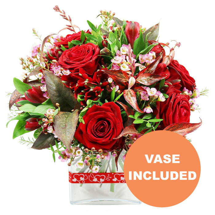 Captured against a clean white backdrop, this romantic arrangement radiates the kind of thoughtful elegance many people in South Norwood look for when sending flowers to mark a milestone or a quiet 'I love you'. The bouquet forms a full, rounded dome of red blooms, centred on several velvety roses, their petals rich and plush, some still tightly furled and others fully open, revealing intricate layers that spiral towards the heart of each flower. Woven between the roses are deep crimson accent flowers resembling alstroemeria or similar multi-flowered stems, their elongated petals and multiple blooms adding intricate texture and a slightly wild, garden-inspired feel. Delicate sprays of tiny white and blush-pink filler flowers, akin to waxflower or limonium, drift through the composition, lending it a soft, airy quality that lightens the intense reds and brings a gentle sense of romance. A lush abundance of greenery frames the bouquet, with wide, glossy leaves and finer, feathery sprigs that suggest freshness and a just-gathered-from-the-garden effect, much like flowers gathered after a stroll past South Norwood Lake. Adding depth and sophistication, the florist has tucked in striking burgundy and reddish-brown leaves, some highlighted with subtle veining, which echo the dark undertones of the roses and introduce a note of refined drama. The flowers are arranged in a clear glass vase with a clean, rectangular base and softly rounded edges, allowing stems and water to be glimpsed within, enhancing the contemporary presentation. Encircling the vase is a festive red ribbon, printed with a simple repeating pattern of white heart outlines, emphasising the bouquet's romantic purpose. In the lower right corner, an orange circular overlay with bold white capital letters announces VASE INCLUDED, clearly indicating this luxurious gift is delivered ready to display in any South Norwood setting.